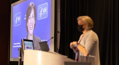 A masked presenter standing at a podium.